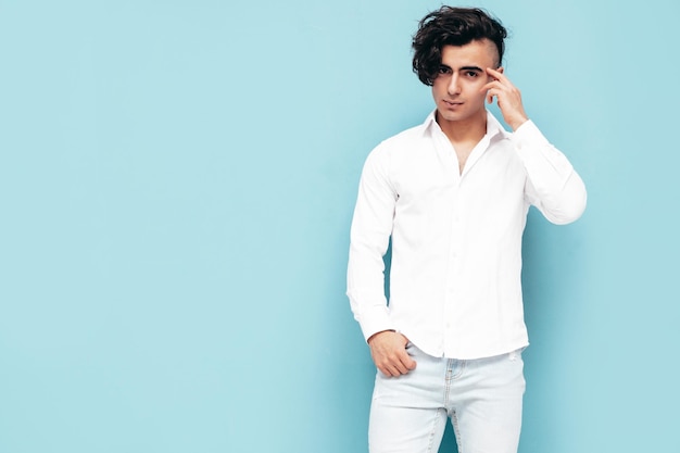 Portrait of handsome confident stylish hipster lambersexual model Sexy man dressed in summer white shirt and jeans clothes Fashion male isolated in studio Posing near blue wall