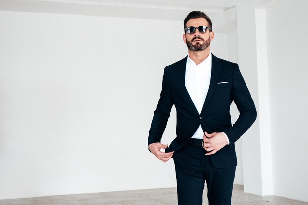 잘 생긴 자신감 세련된 힙스터 램버섹슈얼 모델의 초상화 검은색 우아한 양복을 입은 섹시한 현대 남성 패션 남성 선글라스를 끼고 흰색 배경에 스튜디오에서 포즈를 취합니다.