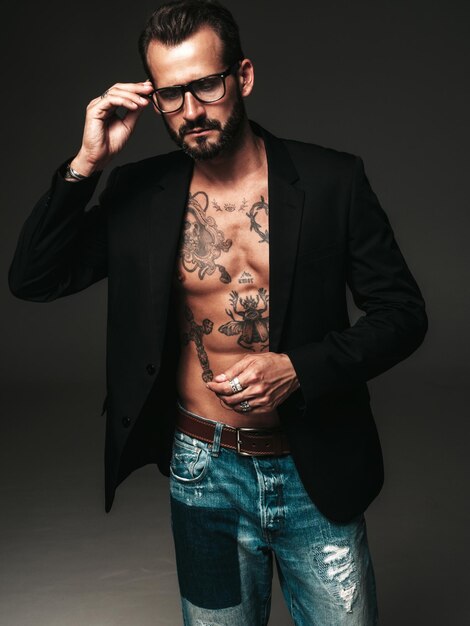 잘 생긴 자신감 세련된 힙스터 램버섹슈얼 모델의 초상화 검은 재킷을 입은 섹시한 현대 남자 문신이 있는 벌거 벗은 몸통 안경에서 어두운 배경에 스튜디오에서 포즈를 취하는 패션 남성