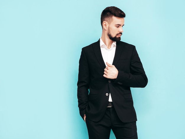 Portrait of handsome confident stylish hipster lambersexual model sexy modern man dressed in elegant black suit fashion male posing in studio near blue wall