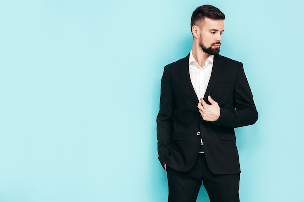 Portrait of handsome confident stylish hipster lambersexual model Sexy modern man dressed in elegant black suit Fashion male posing in studio near blue wall