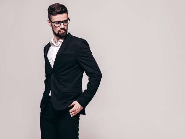 잘 생긴 자신감 세련된 힙스터 램버섹슈얼 모델의 초상화 우아한 검은 양복을 입은 섹시한 현대 남성 안경에 회색으로 격리된 흰 벽 근처 스튜디오에서 포즈를 취하는 패션 남성
