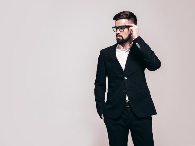 Portrait of handsome confident stylish hipster lambersexual model Sexy modern man dressed in elegant black suit Fashion male posing in studio near white wall Isolated on grey In spectacles