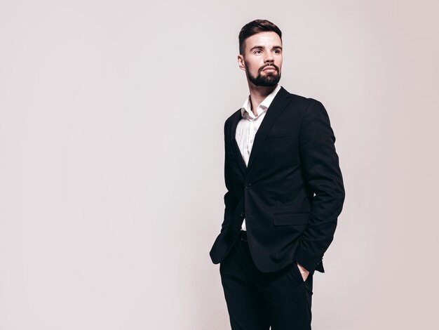 Portrait of handsome confident stylish hipster lambersexual model Sexy modern man dressed in elegant black suit Fashion male posing in studio near white wall Isolated on grey