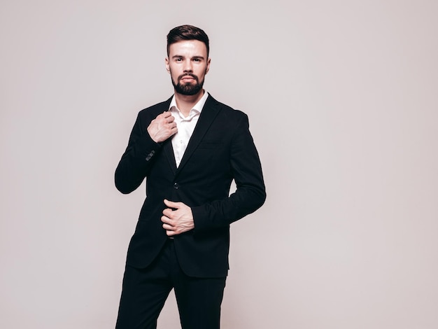 Portrait of handsome confident stylish hipster lambersexual model Sexy modern man dressed in elegant black suit Fashion male posing in studio near white wall Isolated on grey