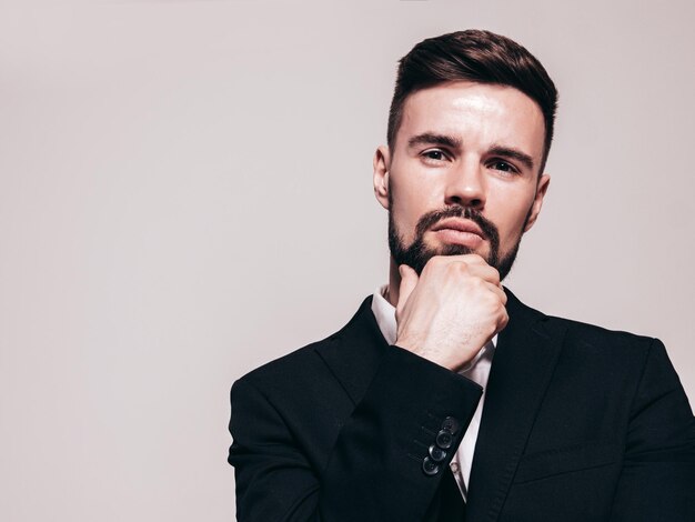 Portrait of handsome confident stylish hipster lambersexual model Sexy modern man dressed in elegant black suit Fashion male posing in studio near white wall Isolated on greyTouching his chin