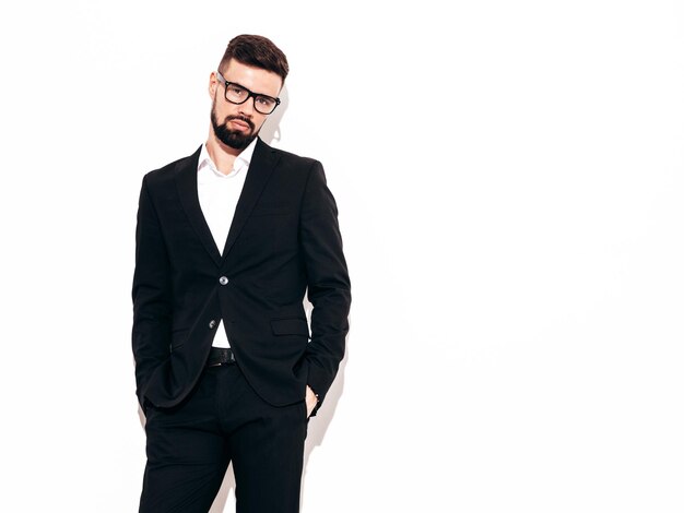 Portrait of handsome confident stylish hipster lambersexual model Sexy modern man dressed in elegant black suit Fashion male posing in studio near white wall Isolated on white In spectacles