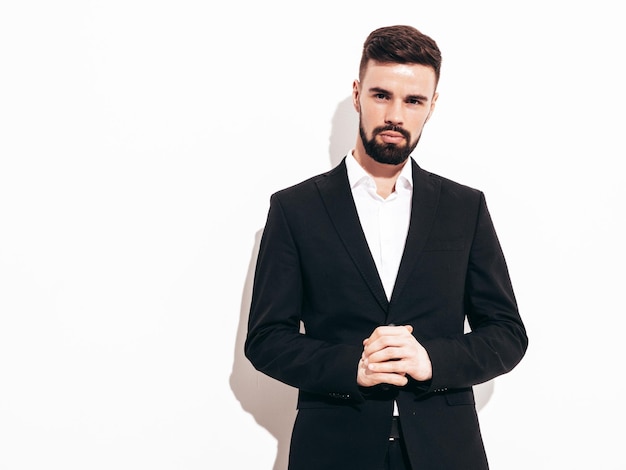 Portrait of handsome confident stylish hipster lambersexual model Sexy modern man dressed in elegant black suit Fashion male posing in studio near white wall Isolated