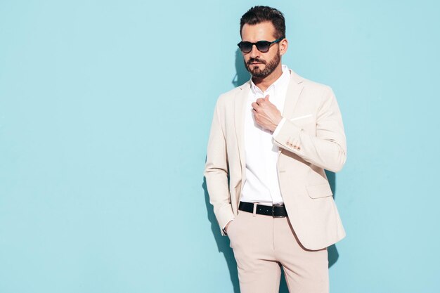 Portrait of handsome confident stylish hipster lambersexual model Sexy modern man dressed in white elegant suit Fashion male posing in studio near blue wall in sunglasses