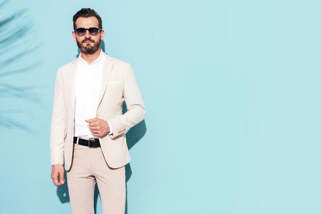잘 생긴 자신감 세련된 힙스터 램버섹슈얼 모델의 초상화 흰색 우아한 정장을 입은 섹시한 현대 남자 패션 남성 선글라스를 끼고 파란색 벽 근처 스튜디오에서 포즈를 취합니다.