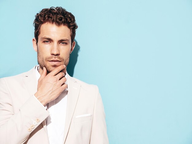 Portrait of handsome confident stylish hipster lambersexual model Sexy modern man dressed in white elegant suit Fashion male posing in studio near blue wall Touching his chin