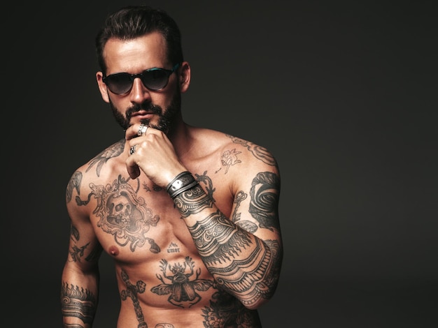 Portrait of handsome confident stylish hipster lambersexual model Sexy modern man Naked torso with tattoosFashion male posing in studio on dark background In sunglasses