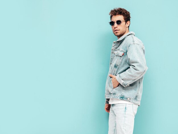 Portrait of handsome confident stylish hipster lambersexual model with curly hairstyle Sexy man dressed in jeans jacket Fashion male isolated on blue wall in studio In sunglasses