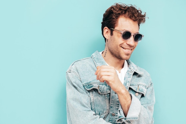 Free photo portrait of handsome confident stylish hipster lambersexual model with curly hairstyle sexy man dressed in jeans jacket fashion male isolated on blue wall in studio in sunglasses