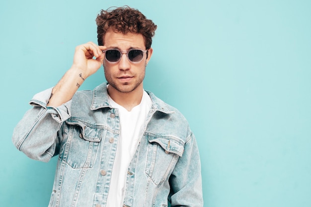 Free photo portrait of handsome confident stylish hipster lambersexual model with curly hairstyle sexy man dressed in jeans jacket fashion male isolated on blue wall in studio in sunglasses