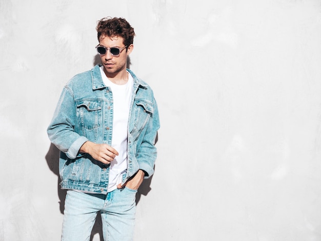 Portrait of handsome confident stylish hipster lambersexual model with curly hairstyle Sexy man dressed in jeans jacket Fashion male isolated on grey wall in studio In sunglasses