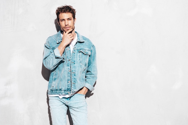 Portrait of handsome confident stylish hipster lambersexual model with curly hairstyle Sexy man dressed in jeans jacket Fashion male isolated on grey wall in studio