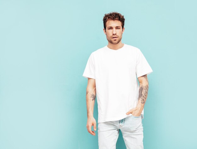 Portrait of handsome confident stylish hipster lambersexual model with curly hairstyle Sexy man dressed in jeans and white tshirt Fashion male isolated on blue wall in studio