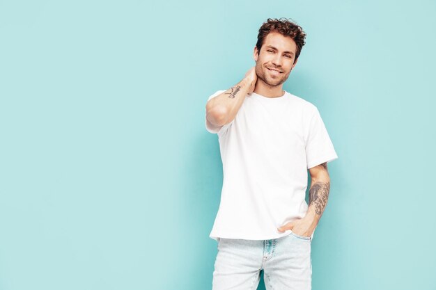 Portrait of handsome confident stylish hipster lambersexual model with curly hairstyle Sexy man dressed in jeans and white tshirt Fashion male isolated on blue wall in studio