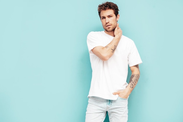 Free photo portrait of handsome confident stylish hipster lambersexual model with curly hairstyle sexy man dressed in jeans and white tshirt fashion male isolated on blue wall in studio