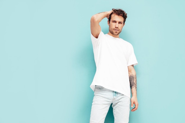 Portrait of handsome confident stylish hipster lambersexual model with curly hairstyle Sexy man dressed in jeans and white tshirt Fashion male isolated on blue wall in studio