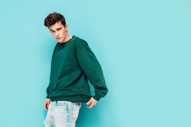 Portrait of handsome confident stylish hipster lambersexual modelMan dressed in green sweater and jeans Fashion male posing in studio near blue wall