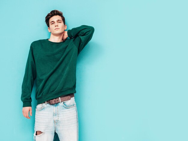 Portrait of handsome confident stylish hipster lambersexual modelMan dressed in green sweater and jeans Fashion male posing in studio near blue wall