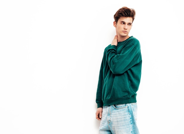 Портрет красивого уверенного в себе стильного хипстера-ламберсексуальной моделиМужчина в зеленом свитере и джинсах Модный мужчина позирует в студии возле белой стены