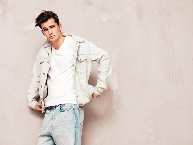 Портрет красивого уверенного в себе стильного хипстера-ламберсексуальной моделиМужчина в куртке и джинсах Модный мужчина позирует в студии возле серой стены