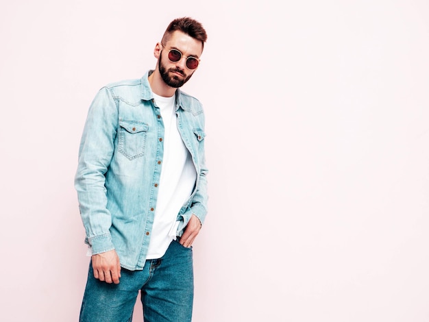 Portrait of handsome confident stylish hipster lambersexual modelMan dressed in jacket and jeans Fashion male posing in studio in sunglasses Isolated on light pink