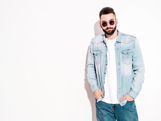 Portrait of handsome confident stylish hipster lambersexual modelMan dressed in jacket and jeans Fashion male posing in studio in sunglasses Isolated on white