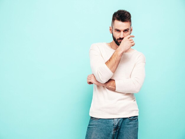 Portrait of handsome confident stylish hipster lambersexual modelMan dressed in white sweater and jeans Fashion male isolated on blue wall in studio Thoughtful