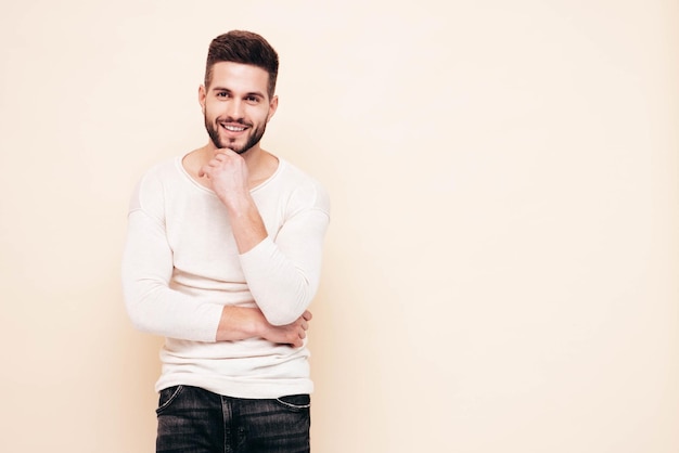 Portrait of handsome confident stylish hipster lambersexual modelMan dressed in white sweater and jeans Fashion male posing near wall in studio