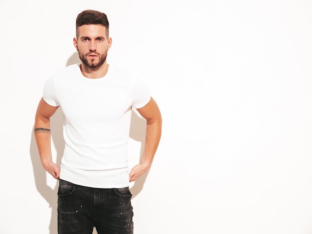 Portrait of handsome confident stylish hipster lambersexual modelMan dressed in white tshirt and jeans Fashion male posing near wall in studio Isolated