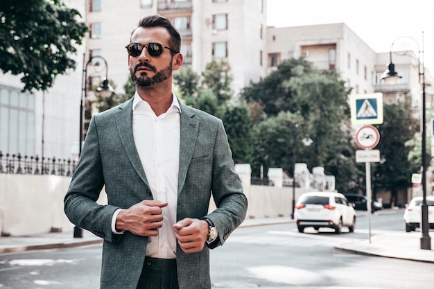 Ritratto di modello lambersexual bello fiducioso elegante hipster uomo moderno vestito con un abito elegante moda maschile in posa sullo sfondo della strada in europa città al tramonto in occhiali da sole