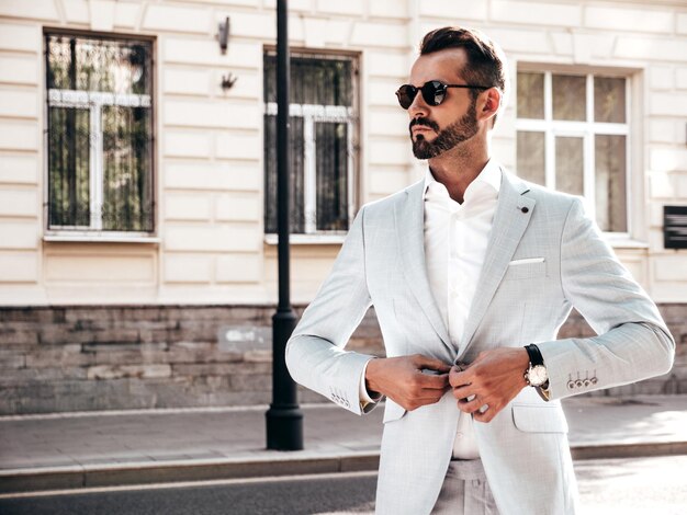 Portrait of handsome confident stylish hipster lambersexual modelModern man dressed in elegant white suit Fashion male posing in the street background in Europe city at sunset In sunglasses