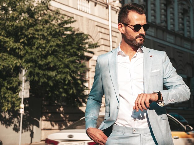Portrait of handsome confident stylish hipster lambersexual modelModern man dressed in elegant white suit Fashion male posing in the street background in Europe city at sunset In sunglasses