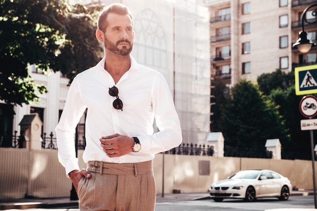 Foto gratuita ritratto di modello lambersexual bello fiducioso elegante hipster uomo moderno vestito con camicia bianca e pantaloni moda maschile in posa sullo sfondo della strada nella città europea al tramonto in occhiali da sole