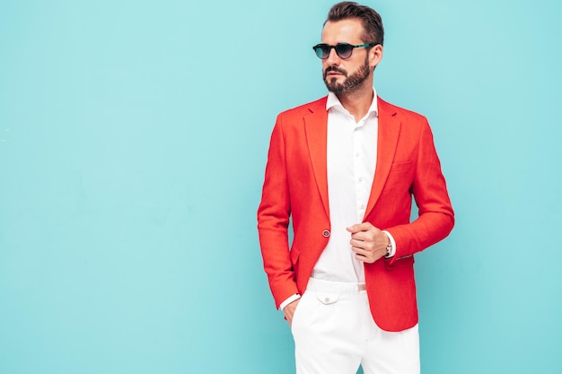 Portrait of handsome confident stylish hipster lambersexual modelSexy modern man dressed in elegant red suit Fashion male posing in studio near blue wall in sunglasses