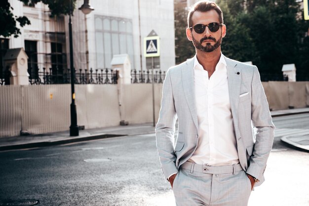 Portrait of handsome confident stylish hipster lambersexual modelSexy modern man dressed in elegant white suit Fashion male posing in the street background in Europe city at sunset In sunglasses