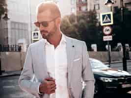 Free photo portrait of handsome confident stylish hipster lambersexual modelsexy modern man dressed in elegant white suit fashion male posing in the street background in europe city at sunset in sunglasses