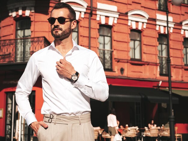 Portrait of handsome confident stylish hipster lambersexual modelSexy modern man dressed in white shirt and trousers Fashion male posing on street background in Europe city at sunset In sunglasses