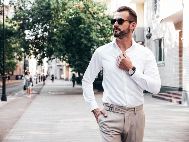 Portrait of handsome confident stylish hipster lambersexual modelSexy modern man dressed in white shirt and trousers Fashion male posing on street background in Europe city at sunset In sunglasses