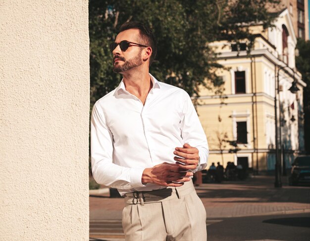 Portrait of handsome confident stylish hipster lambersexual modelSexy modern man dressed in white shirt and trousers Fashion male posing on street background in Europe city at sunset In sunglasses