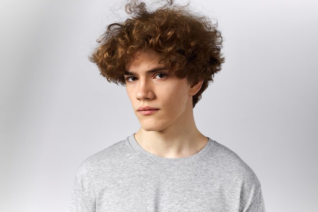Portrait of handsome cool teenage boy with voluminous hair, brown eyes and smooth face dressed in casual gray t-shirt staring at camera with serious confident look. Skin care and youth concept