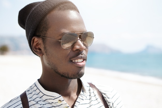 Portrait of handsome dark-skinned hipster in mirrored shades and stylish hat, looking at distance with bag on his shoulders