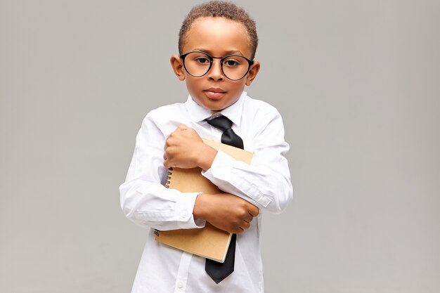 Portrait of handsome dark skinned schoolboy with short Afro haircut posing isolated in glasses, shirt and tie embracing copybook, feeling shy in new school. Learning and knowledge