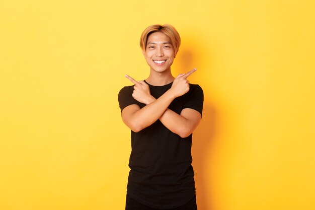Portrait of handsome determined blond asian guy, student pointing fingers sideways, showing two variants, standing yellow wall.