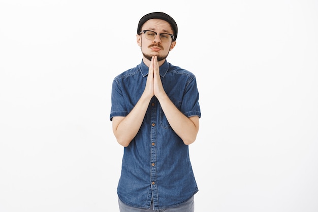 Portrait of handsome and emotive handsome bearded man in glasses and trendy black beanie holding hands in pray near chin squinting and tilting head asking forgiveness or favor