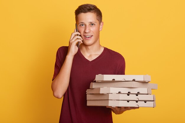  portrait of handsome fair haired young teenager holding smartphone and boxes with food, bringing order, phoning client